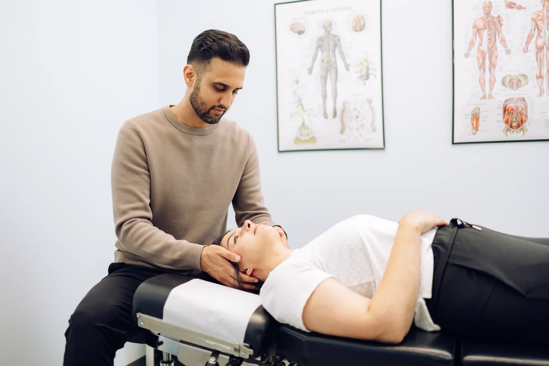 A man in a beige sweater is performing a neck adjustment on a woman lying on a treatment table in a clinic, with anatomical charts on the wall in the background.