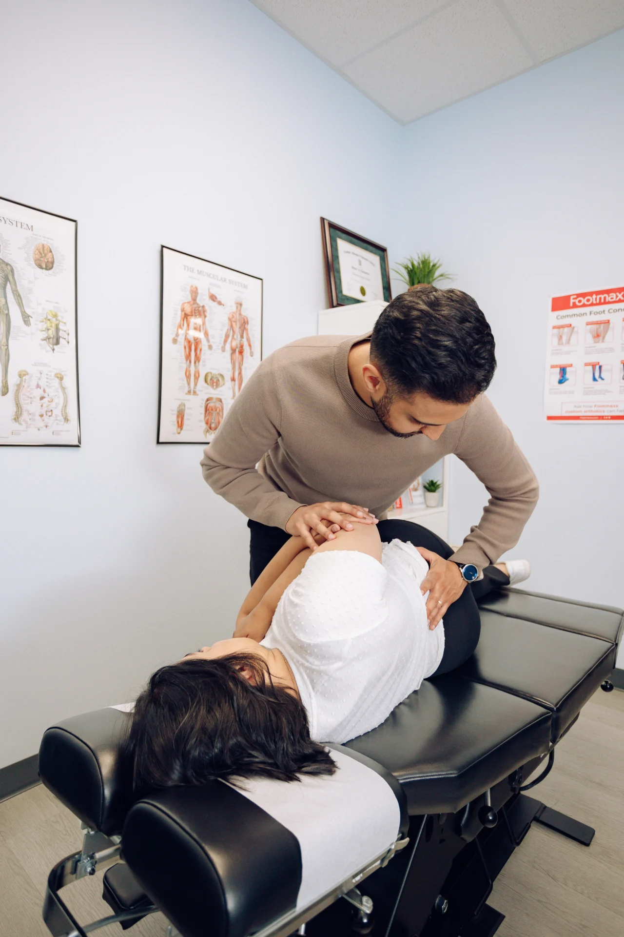 A chiropractor adjusts a woman's back as she lies on a table in a treatment room with anatomical posters on the walls.