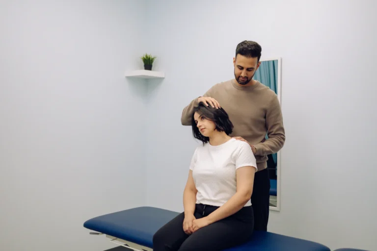A person in a white shirt is sitting on a blue table in a medical office, while another person in a brown sweater stands behind, examining their neck. A mirror and a plant are on the wall.