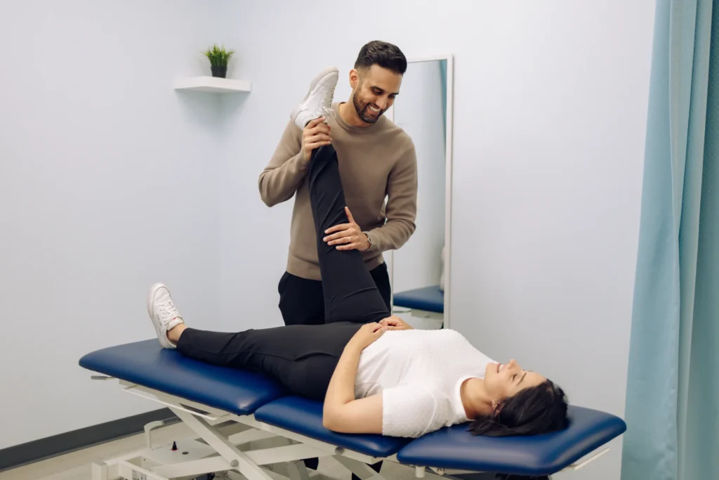 A man assists a woman lying on a treatment table with a leg stretch in a therapy room.