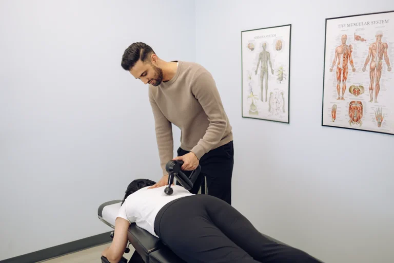 A person receives a massage therapy session using a massage gun in a clinical room with anatomical posters on the wall.