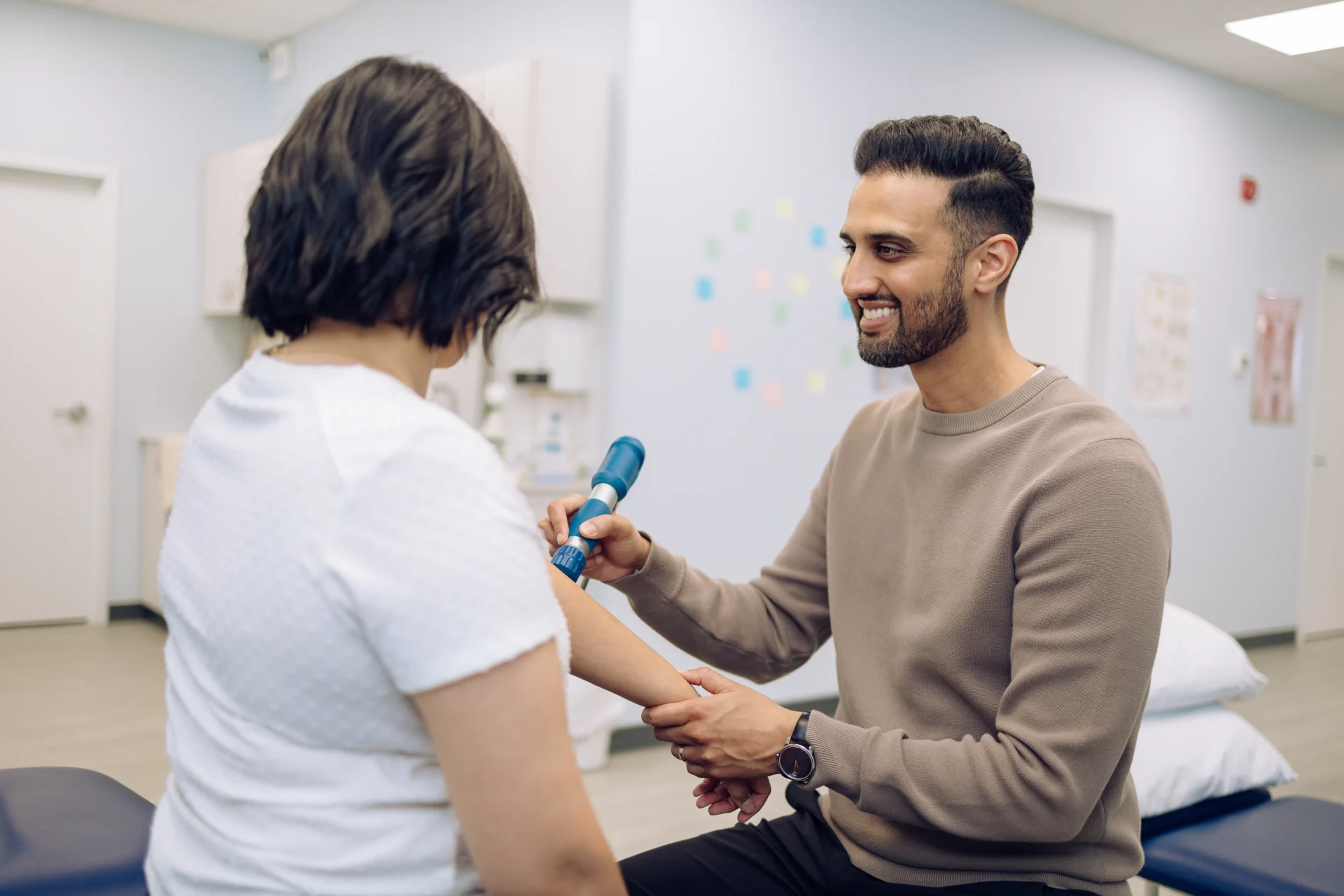 A person in a white shirt receives a treatment from a person using a handheld device in a clinical setting.