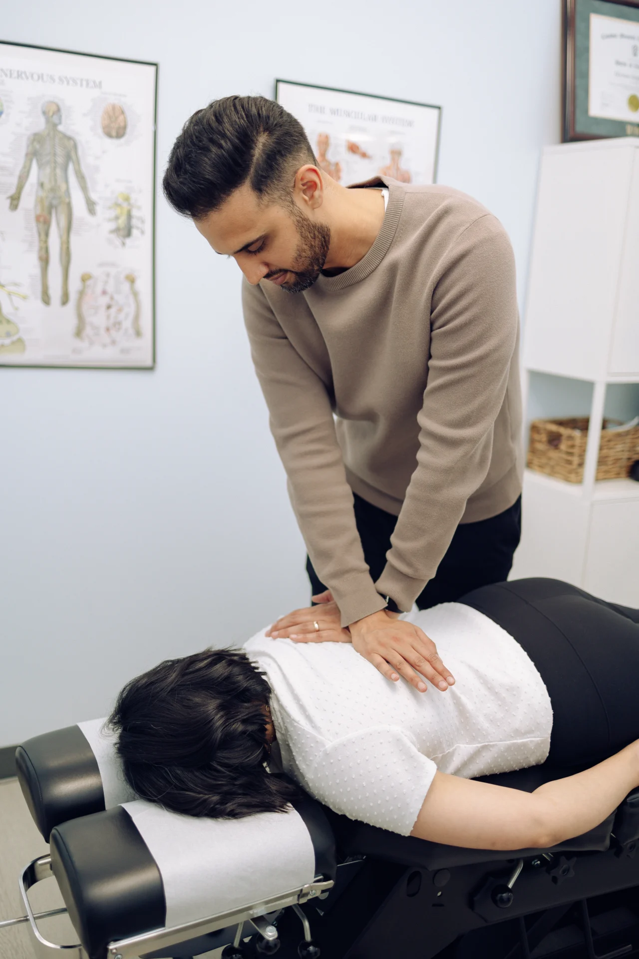 Dr-Harman-Chiropractic-adjustment-1 - Dr. Harman Braich Chiropractic A person in a white shirt lies face down on a chiropractic table while a man performs an adjustment on their back in a clinic.