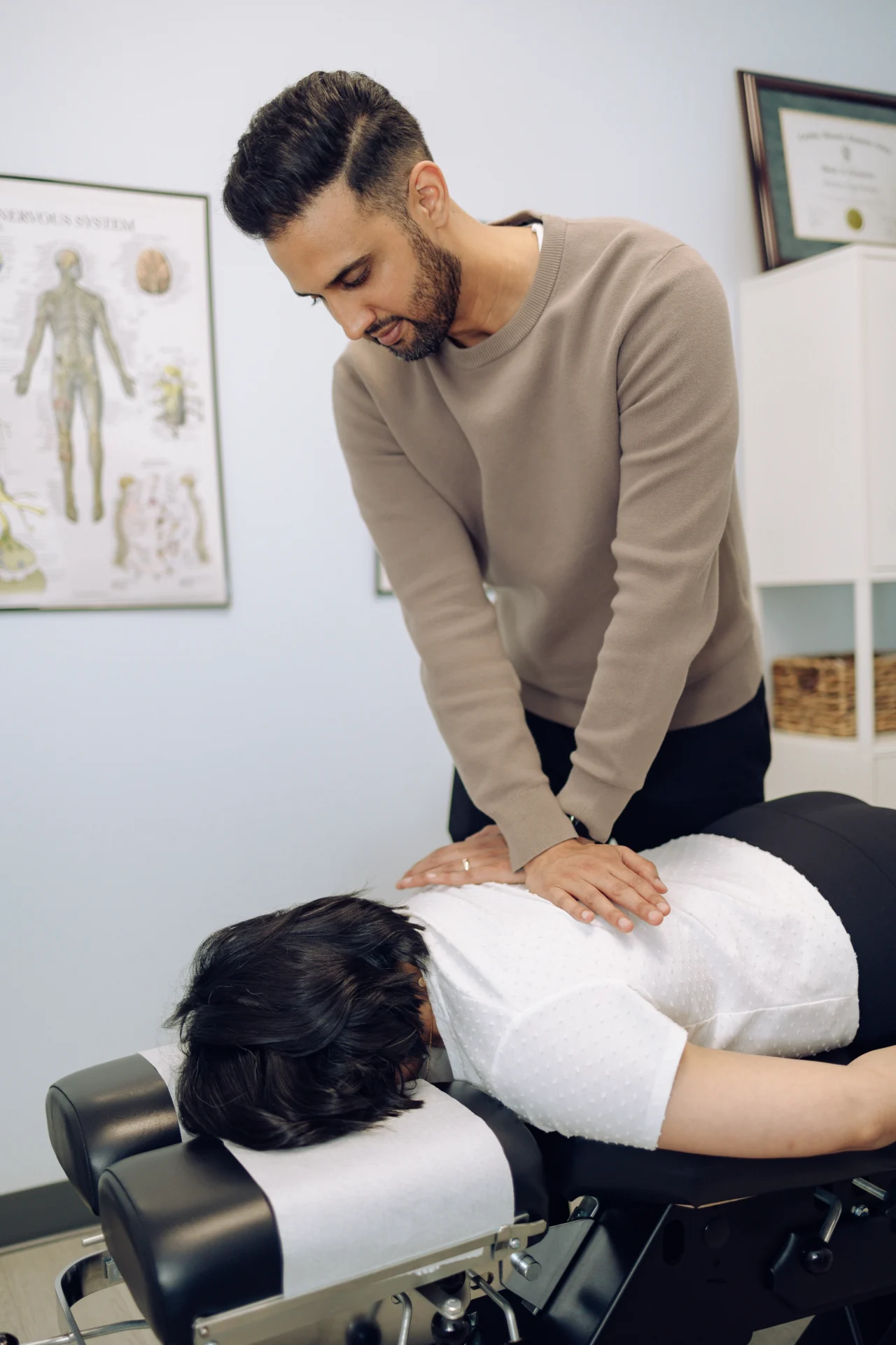 A chiropractor is adjusting a patient's back in a clinic. Medical charts and certificates are visible on the wall.