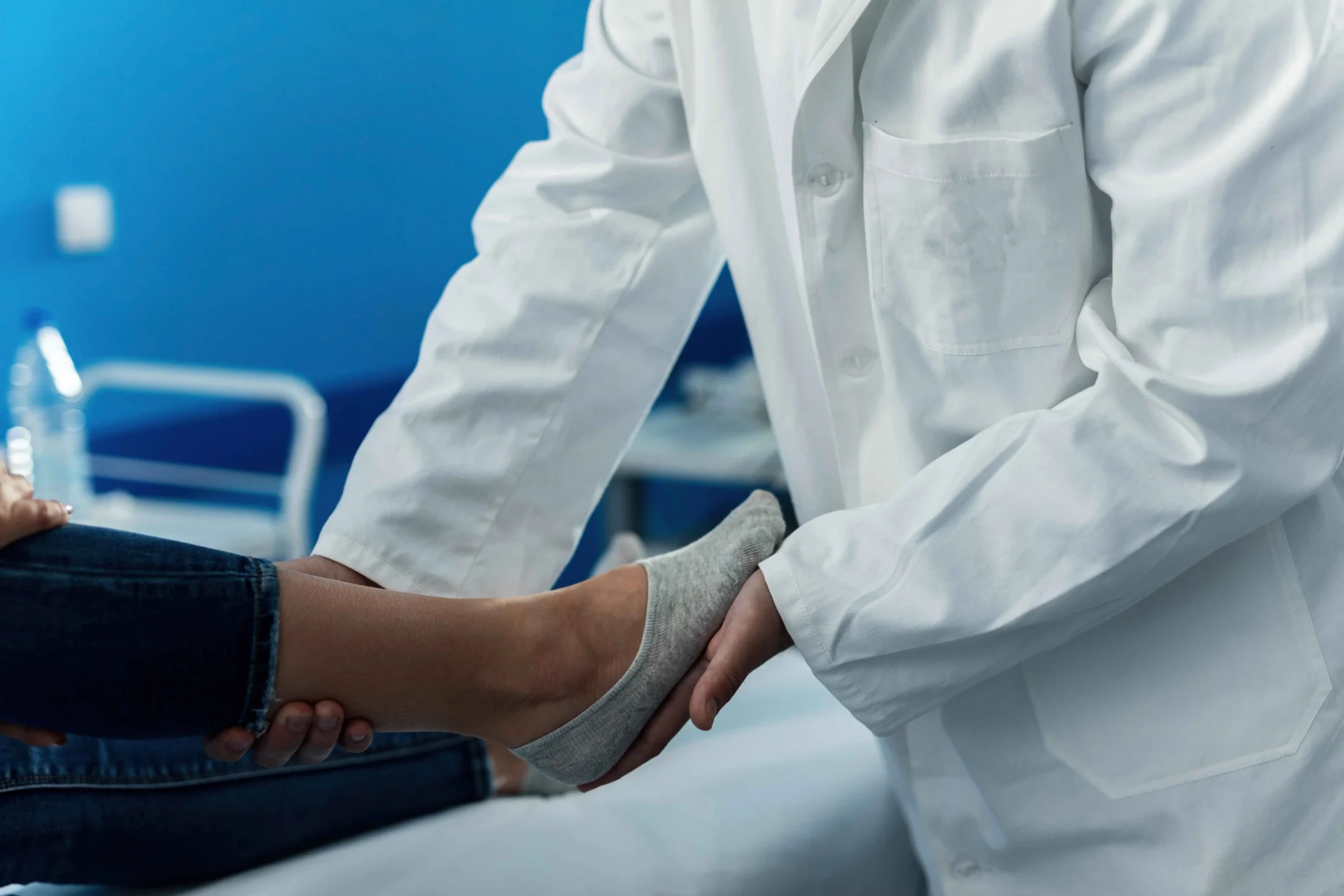 A doctor in a white coat examines a patient's foot, holding it with both hands in a medical setting.