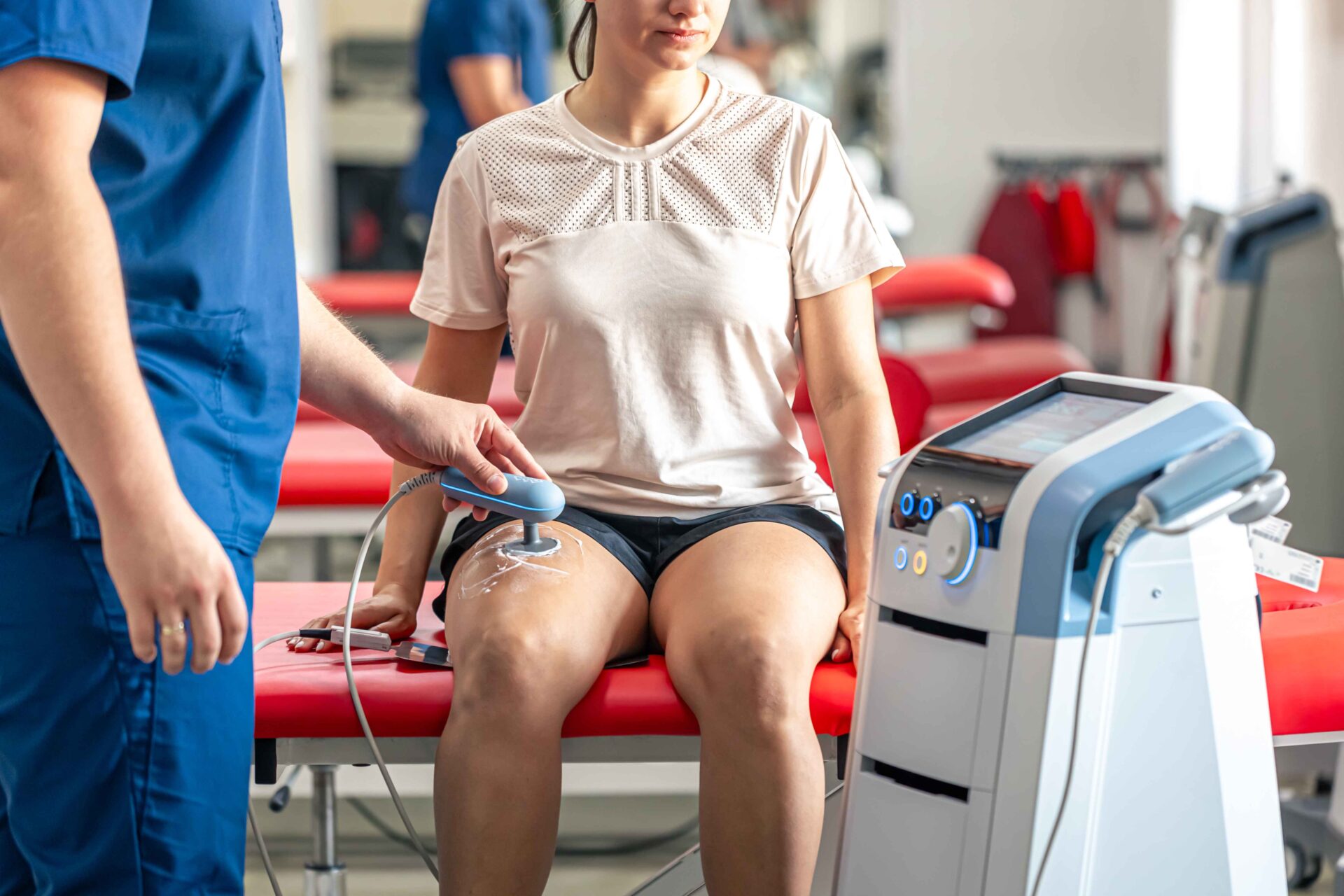 Physical therapist uses an ultrasound therapy device on a patient's knee while seated on a red table.