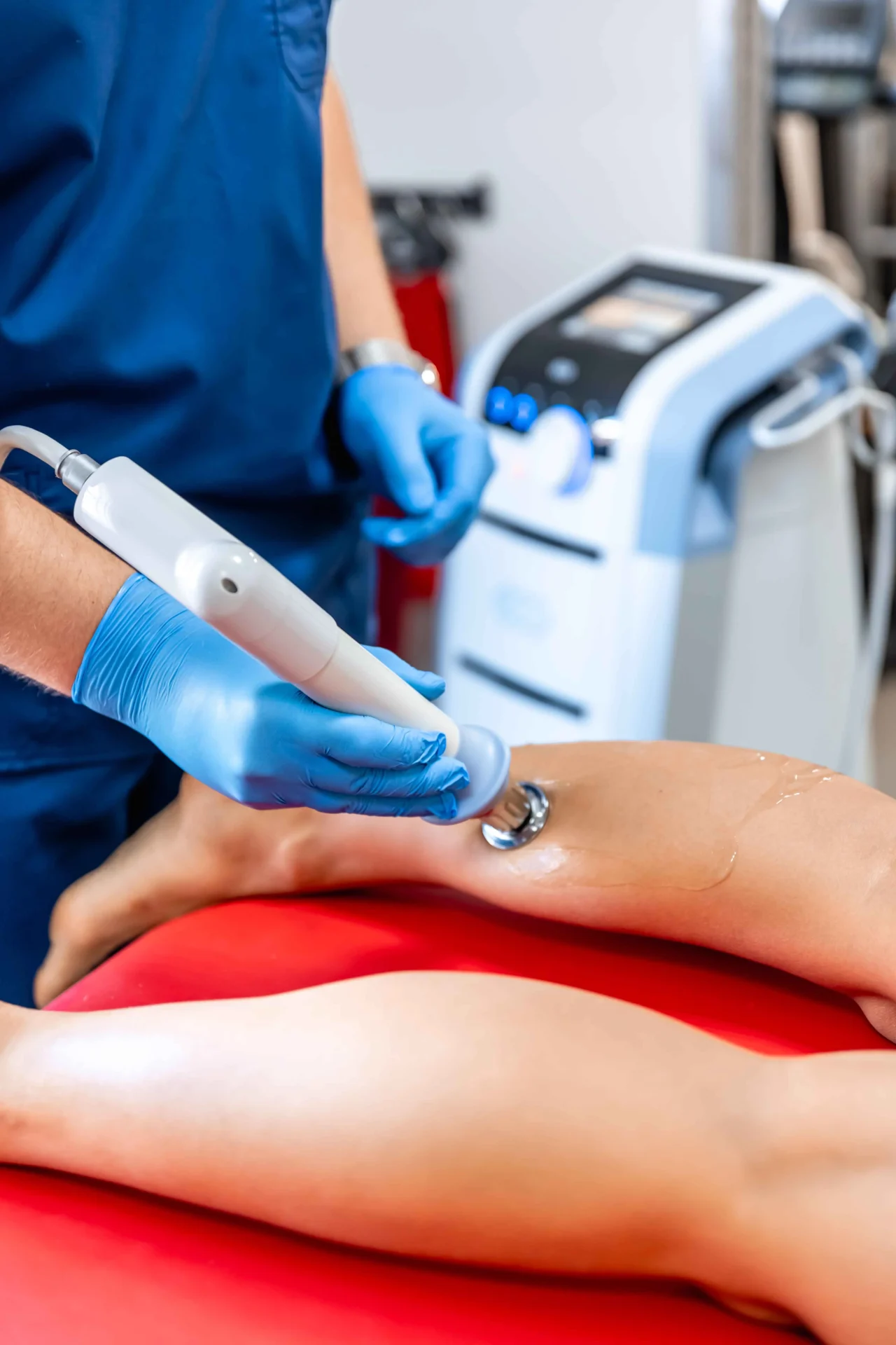 Person receiving a leg treatment with a handheld device operated by a professional wearing blue gloves and scrubs, near a medical machine.