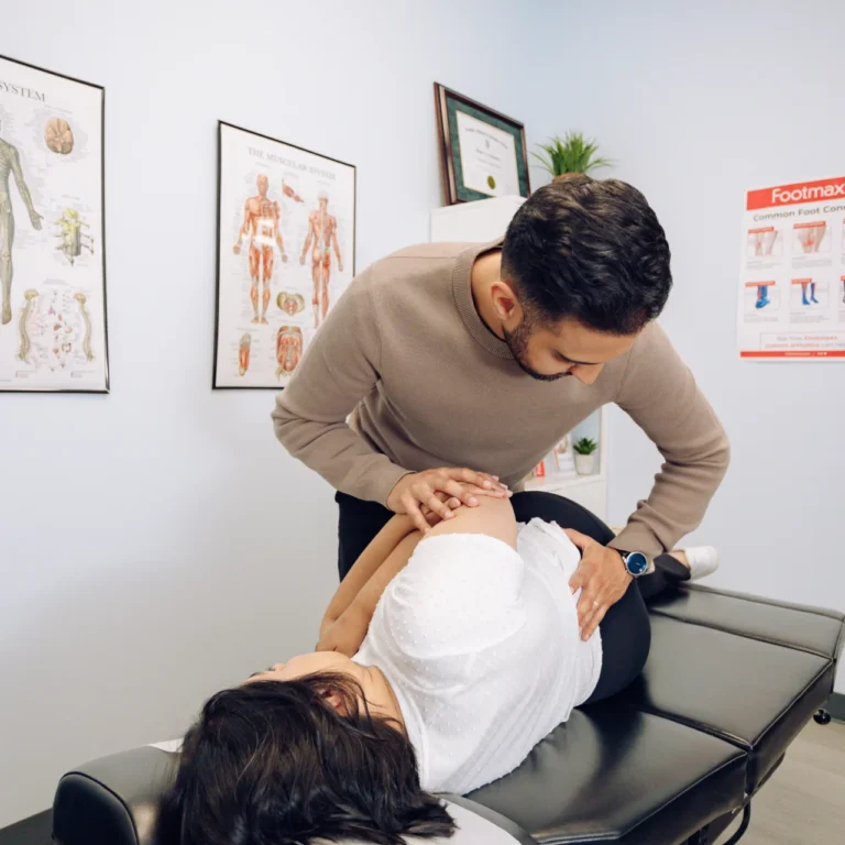 A chiropractor adjusts a woman's back as she lies on a table in a treatment room with anatomical posters on the walls.